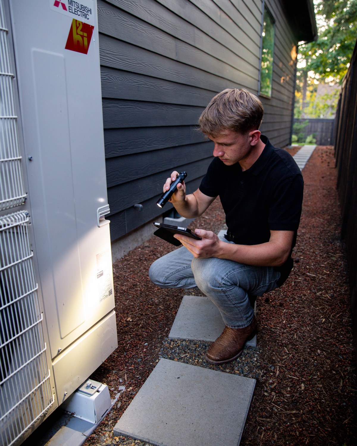 Sam Belza examining exterior heating and cooling equipment beside a home